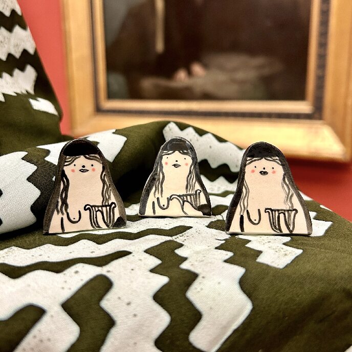 Three Alex Sickling Sappho pin badges displayed in front of Sappho Painting the gallery