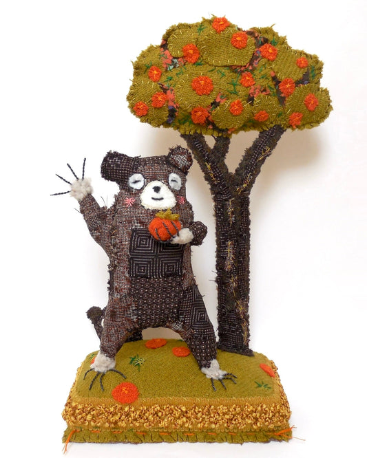 Hand stitched textile sculpture of a brown bear holding an orange persimmon. The bear is stood on a green cloth plinth next to a brown, green and orange persimmon tree. The bear is waving and smiling..