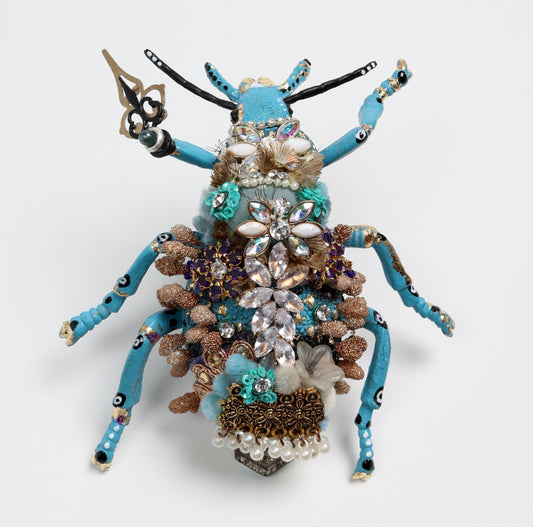 Handmade sculptural beetle figure with a turquoise blue body, long curved legs, and raised antennae, richly decorated with crystals, pearls, beads, feathers, and floral jeweled embellishments, set against a clean white background.