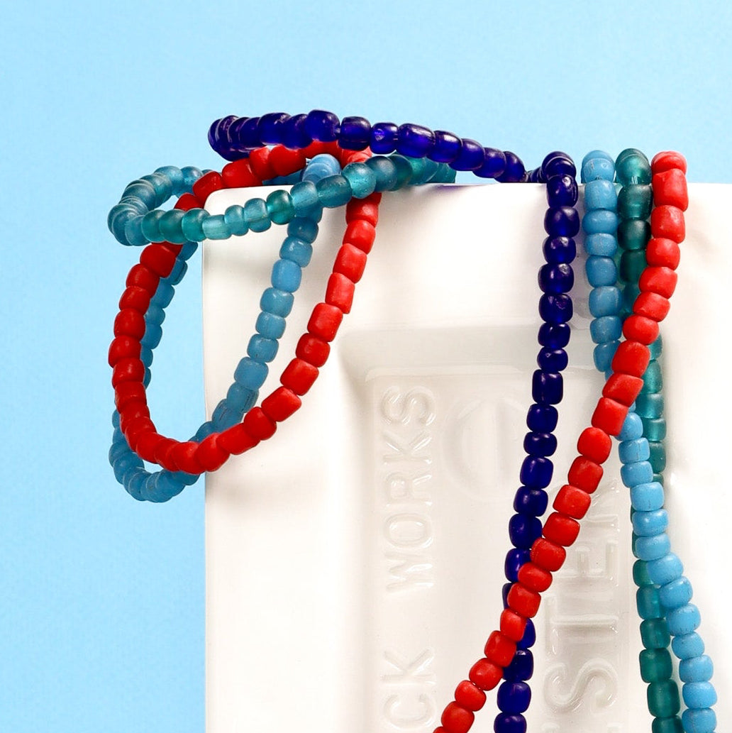 Four beaded bracelets in red, blue, and green draped over a white brick on a light blue background.