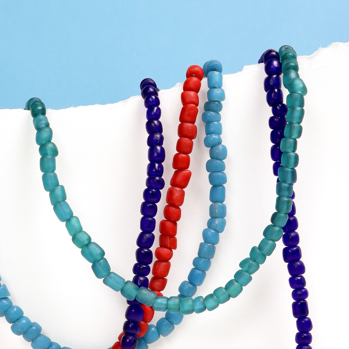 Three beaded necklaces in red, blue, and green on a white surface with a light blue background.