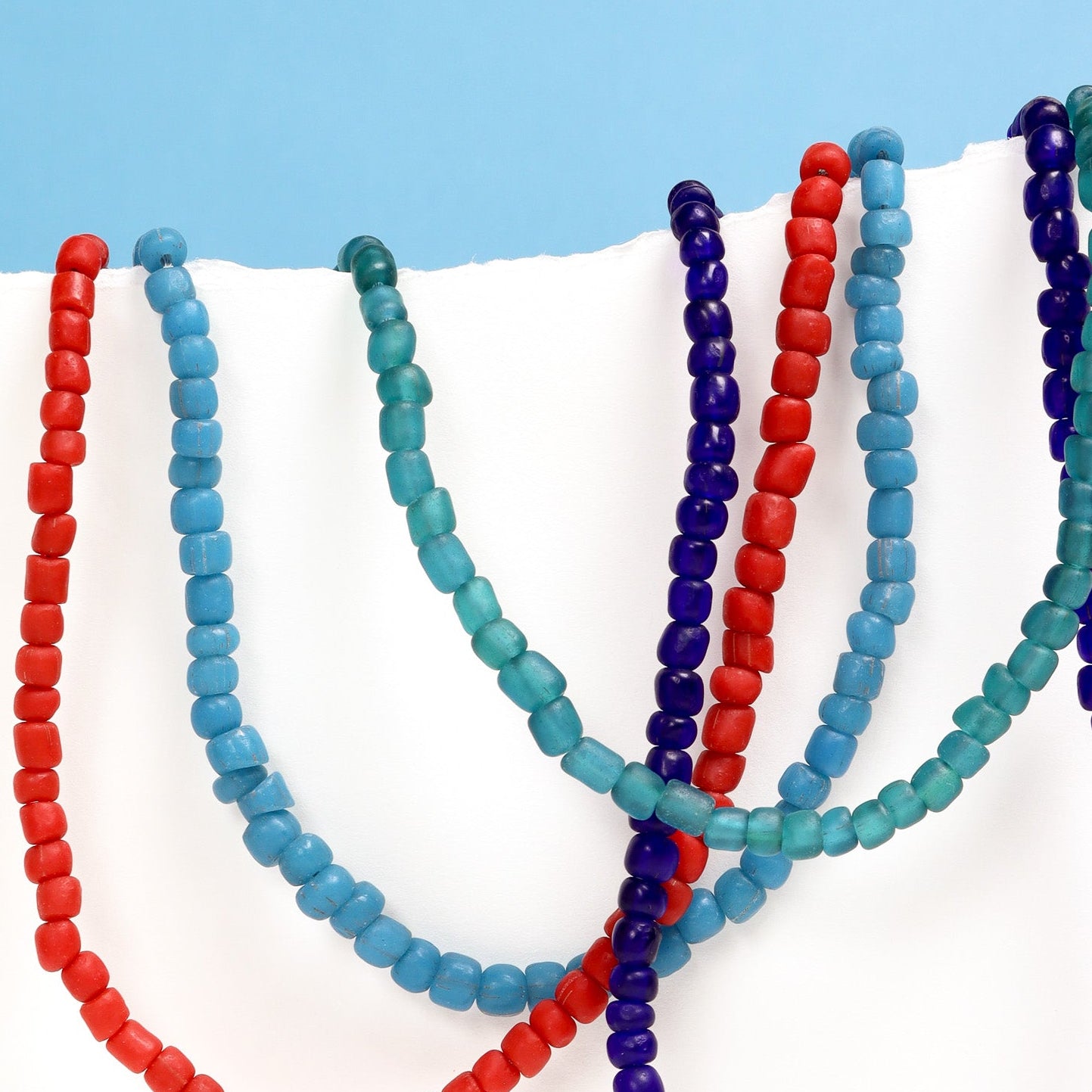 Four beaded necklaces in red, dark blue, light blue and green on a white surface with a light blue background.